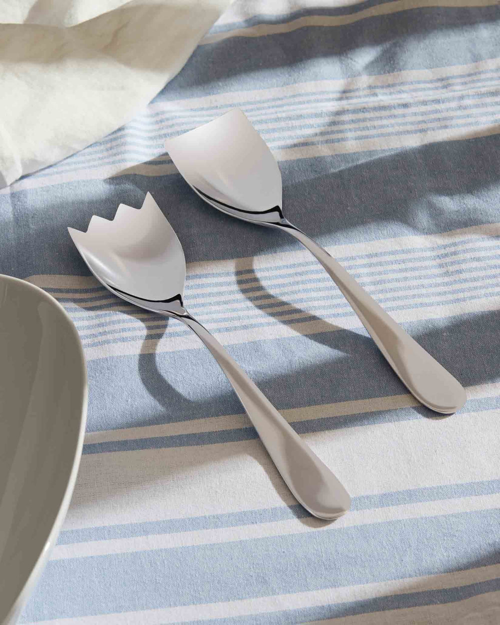 Silver salad servers by Alessi with a contemporary design, one featuring a forked edge, on a blue and white striped tablecloth, next to a white bowl.