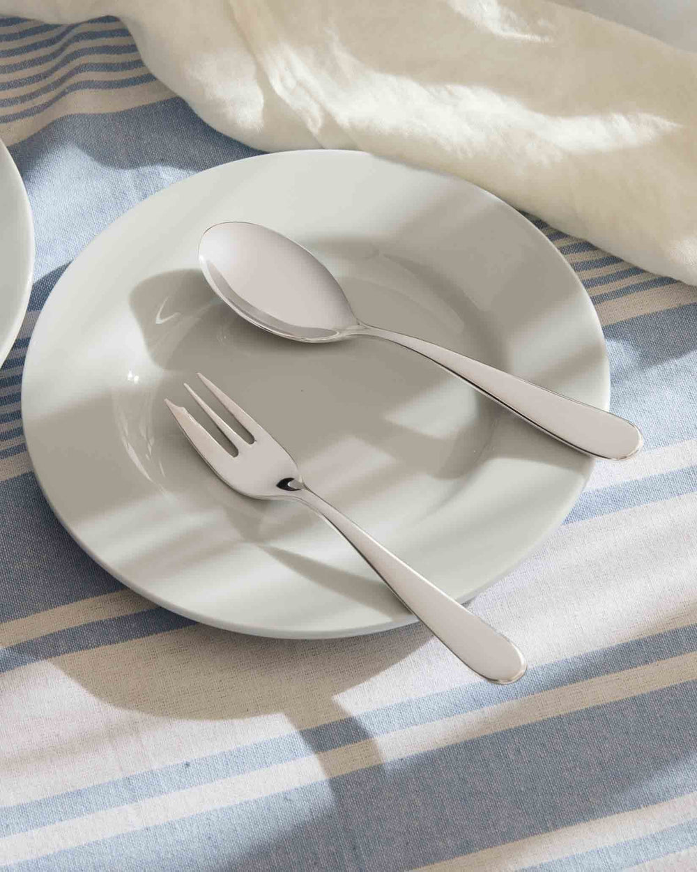 Sleek stainless steel spoon and fork by Alessi on a white plate and striped tablecloth.