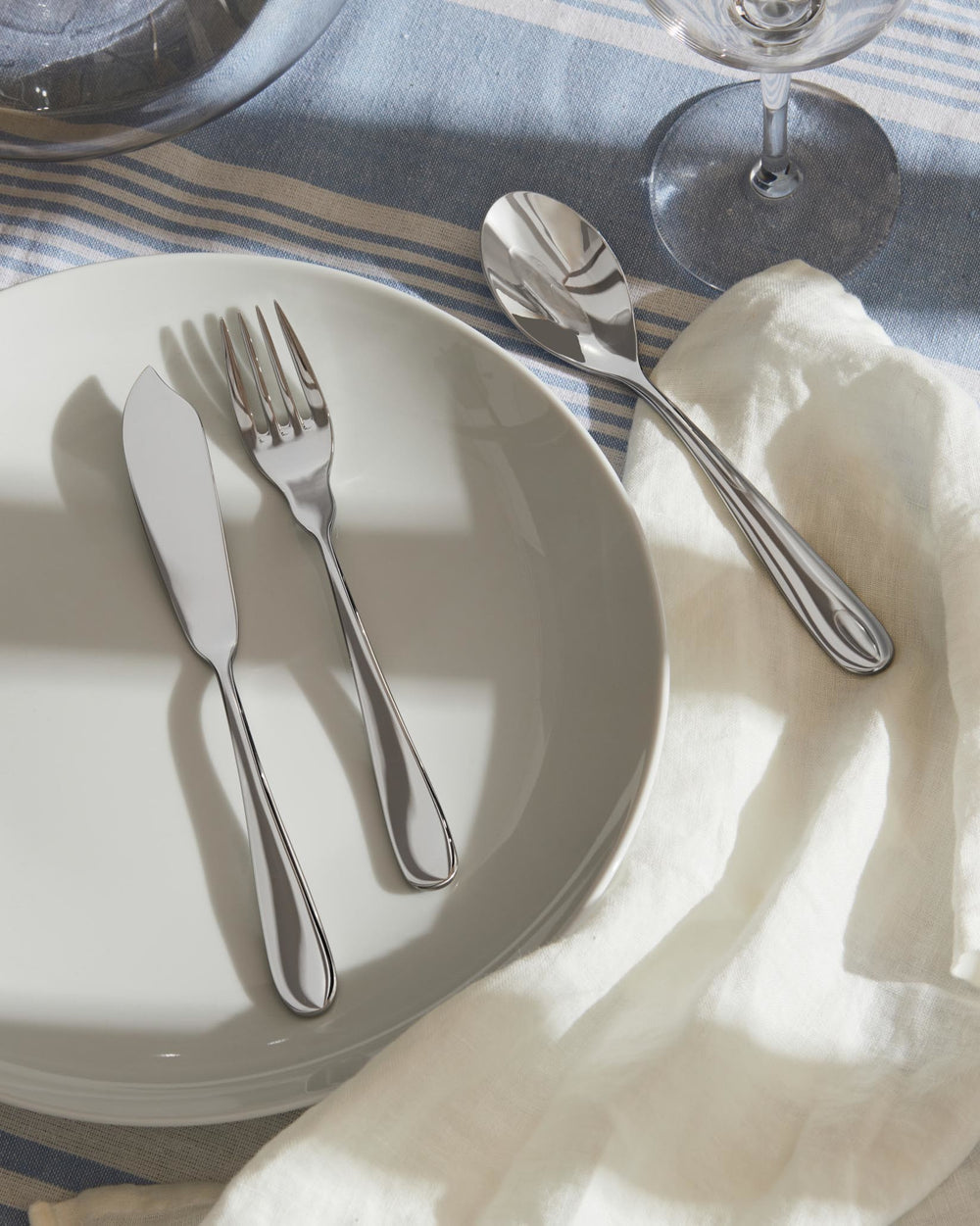 Polished silver butter knife, fork, and spoon by Alessi, neatly arranged on a white plate, with a striped blue tablecloth and white napkin.