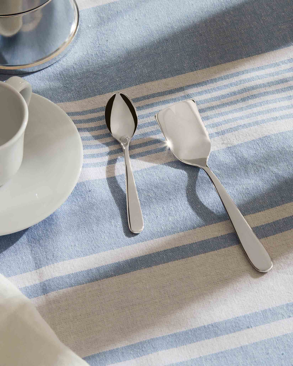 Sleek stainless steel cutlery pieces by Alessi, featuring a spoon and flat serving piece on a blue and white striped tablecloth.