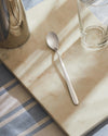 Sleek silver stainless steel spoon by Alessi on a light marble board with a clear glass and striped tablecloth.
