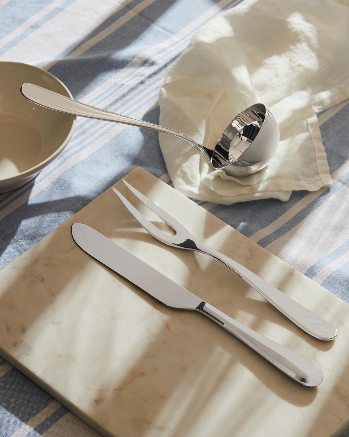 Sleek silver-toned serving ladle, carving knife, and fork by Alessi on a marble board and striped tablecloth.