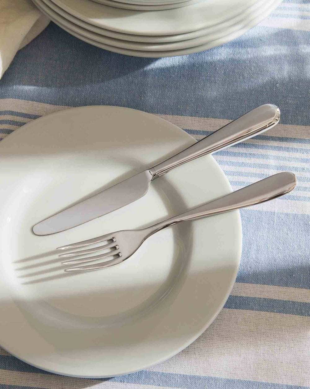 Silver Nuovo Milano fork and knife by Alessi, resting on a white plate on a blue and white striped tablecloth.