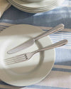 Silver fork and knife by Alessi resting on a white plate on a blue and white striped tablecloth.