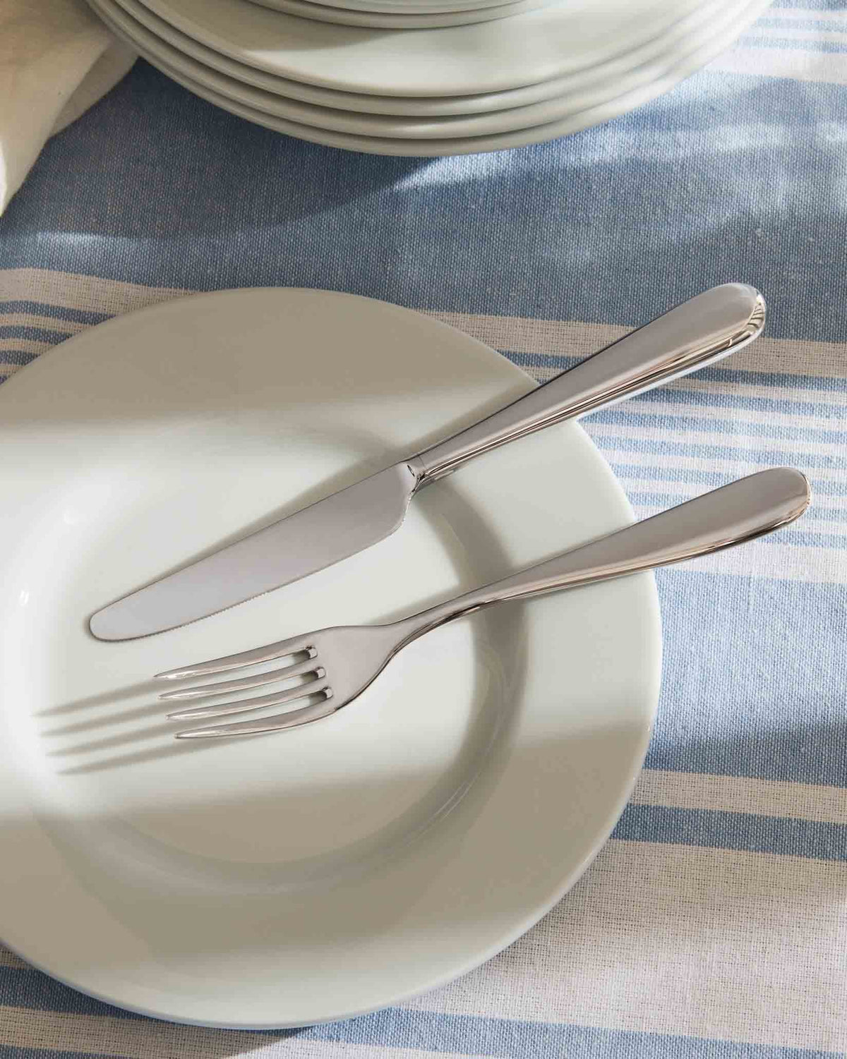 Silver fork and knife by Alessi resting on a white plate on a blue and white striped tablecloth.