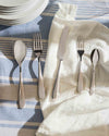 Polished stainless steel cutlery set by Alessi on a striped blue and white tablecloth with a white napkin.
