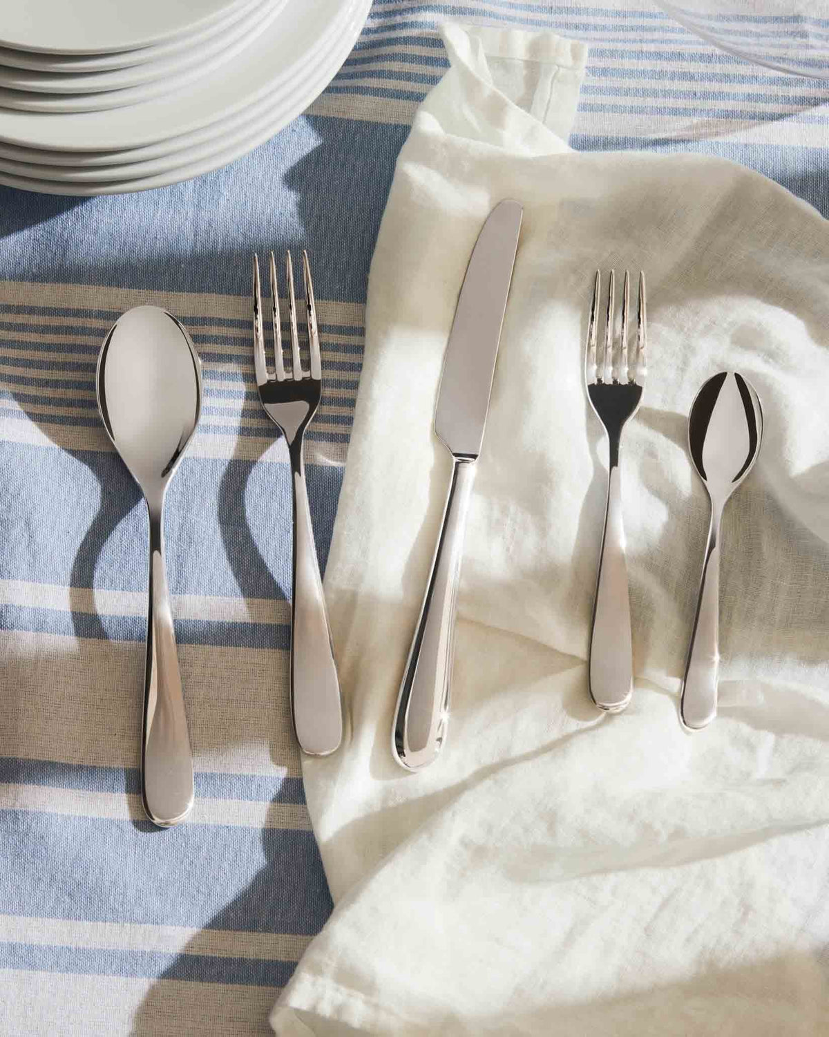 Polished stainless steel cutlery set by Alessi on a striped blue and white tablecloth with a white napkin.