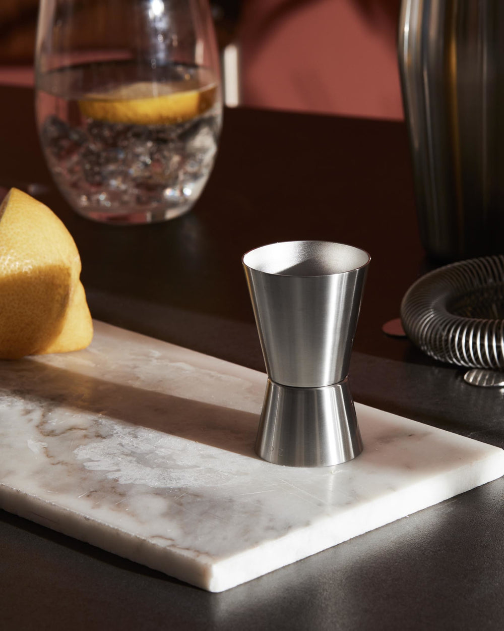 Brushed stainless steel double jigger by Alessi, standing on a white marble slab next to a lemon and a glass of water.