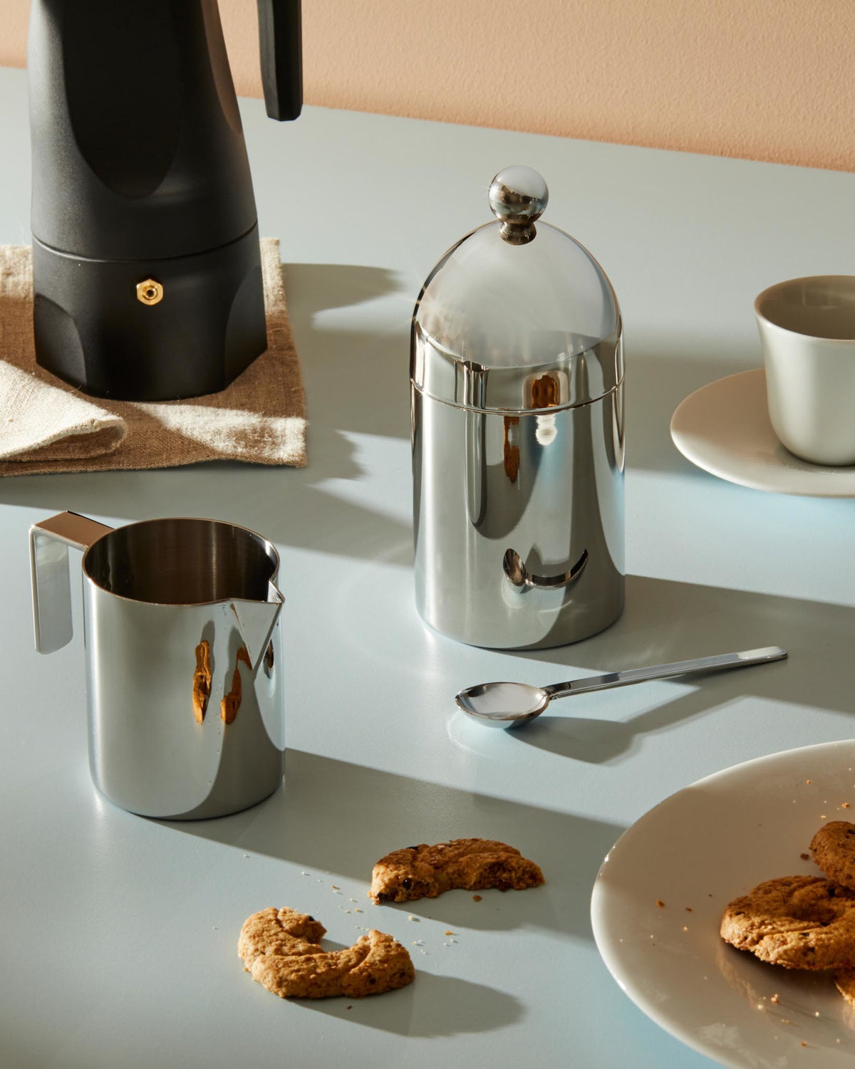 Polished stainless steel sugar bowl by Alessi with a domed lid and spherical knob handle on a light blue table.