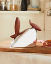 Silver kettle by Alessi with two dark brown wooden handles, on a cutting board with pretzels in a bright kitchen.