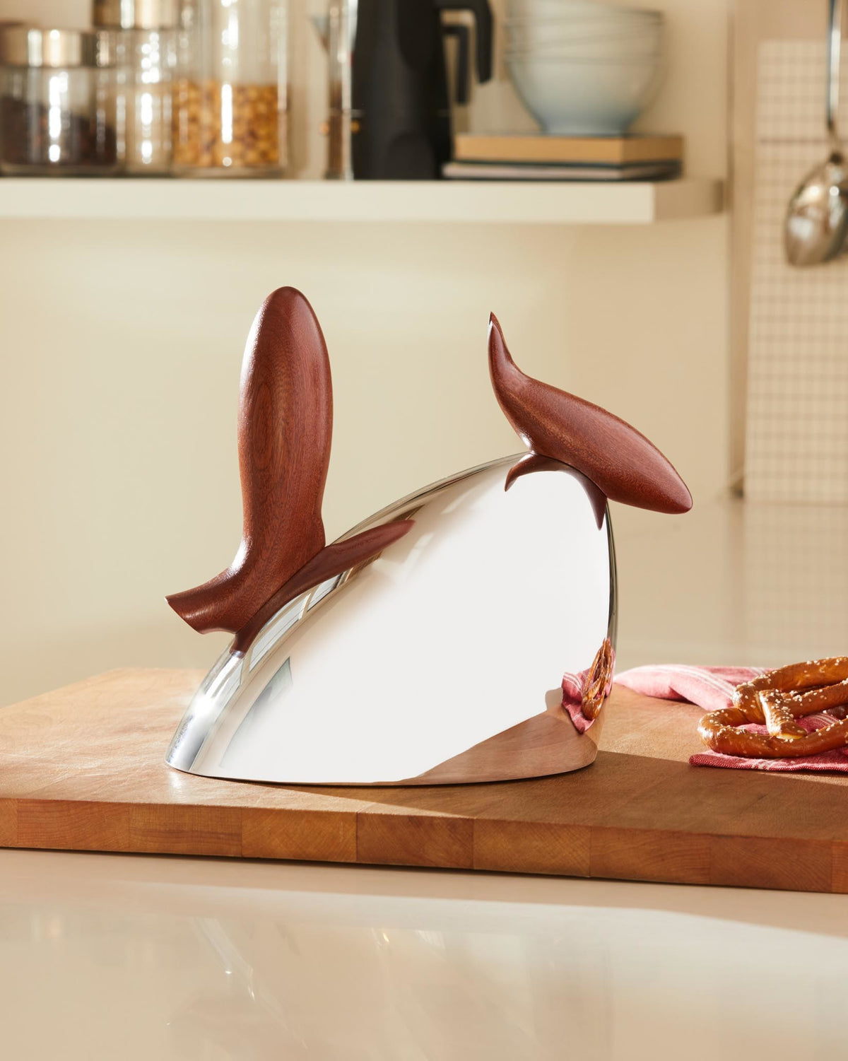Silver kettle by Alessi with two dark brown wooden handles, on a cutting board with pretzels in a bright kitchen.