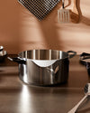 Polished stainless steel cooking pot by Alessi, featuring a curved front rim and black handles on a kitchen counter.