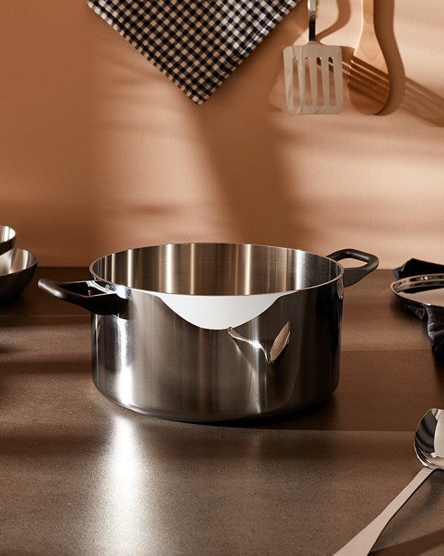 Polished stainless steel cooking pot by Alessi, featuring a curved front rim and black handles on a kitchen counter.
