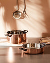 Polished copper pot and shallow pan by Alessi with black handles, reflecting light on a counter.