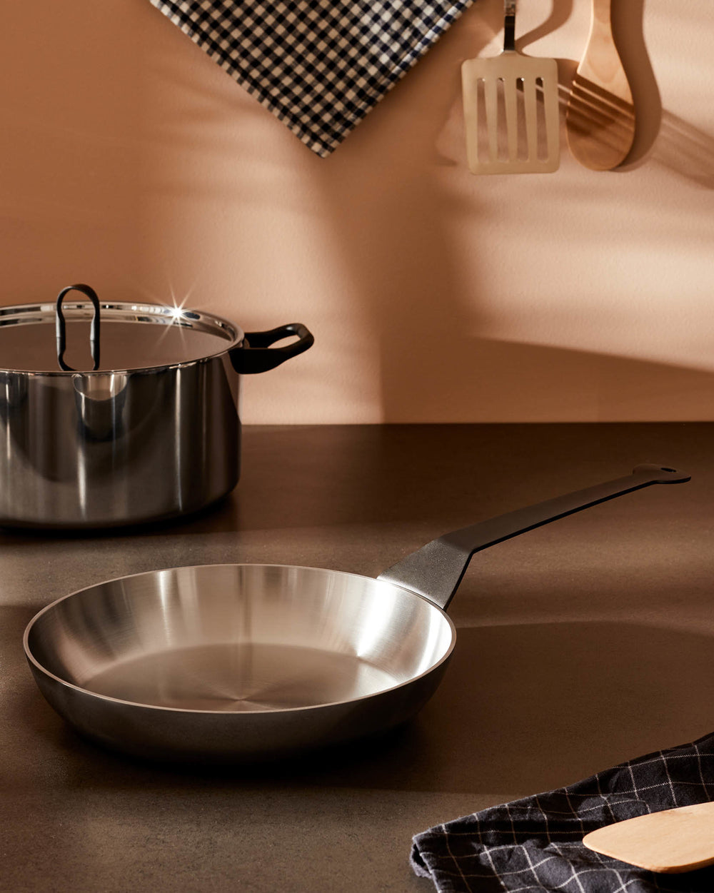 Stainless steel frying pan and cooking pot by Alessi, on a brown counter with utensils and a checkered towel.
