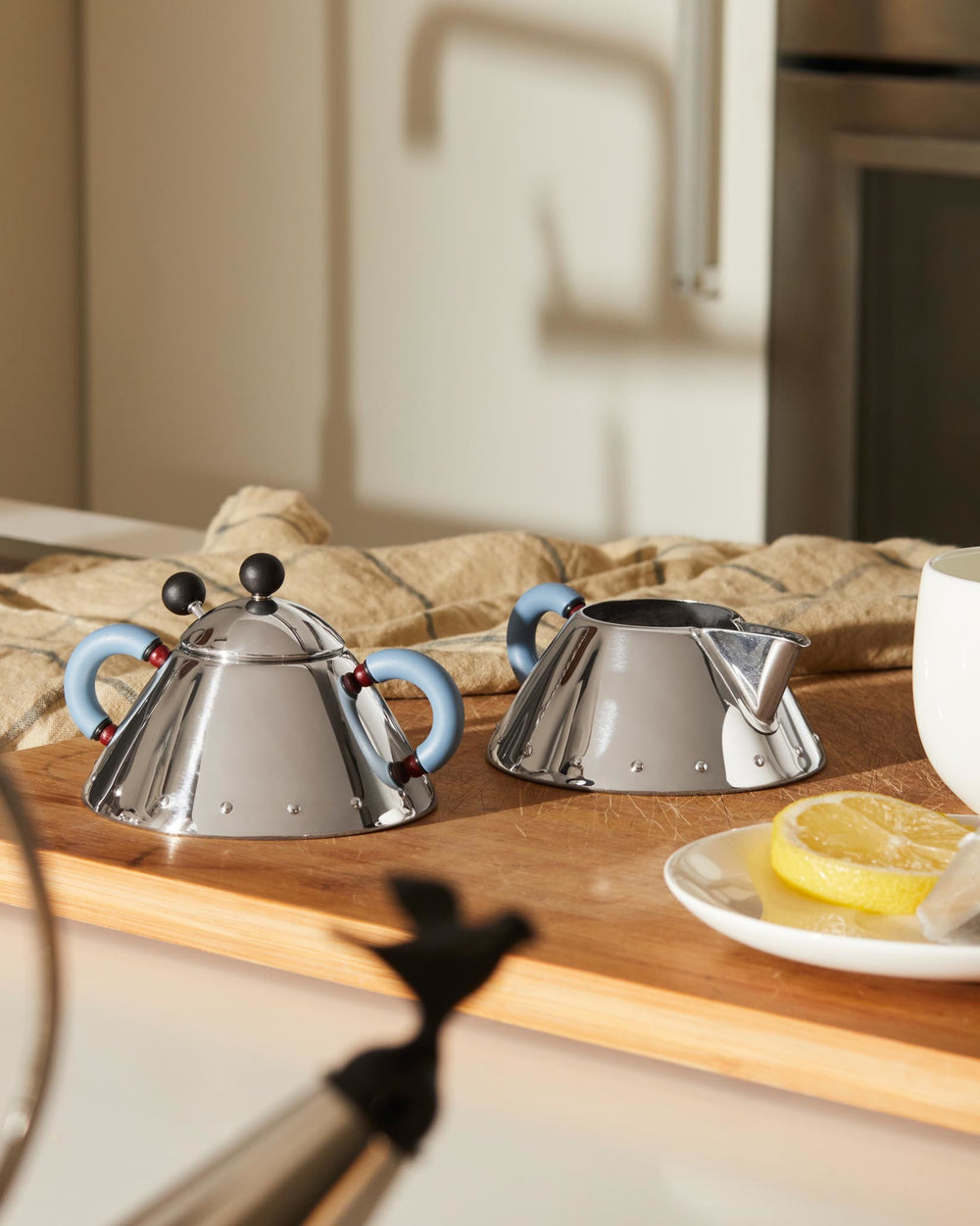 Stainless steel conical tea set by Alessi, featuring a teapot with light blue handles and black finials, and a creamer on a wooden board.