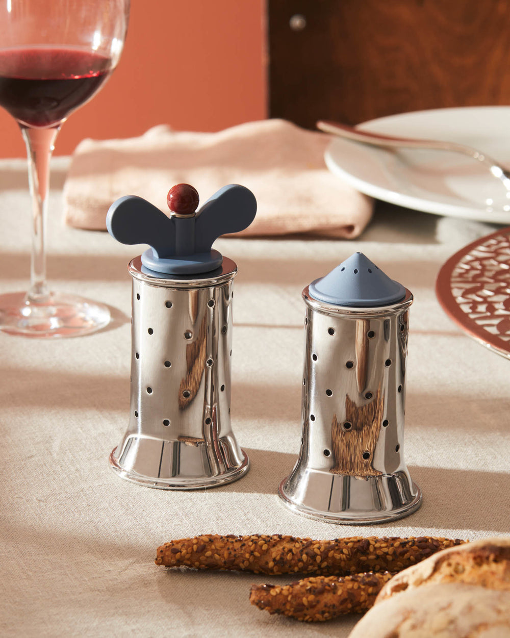 Silver stainless steel perforated shakers by Alessi, one with a blue butterfly top and red knob, the other with a blue conical top.