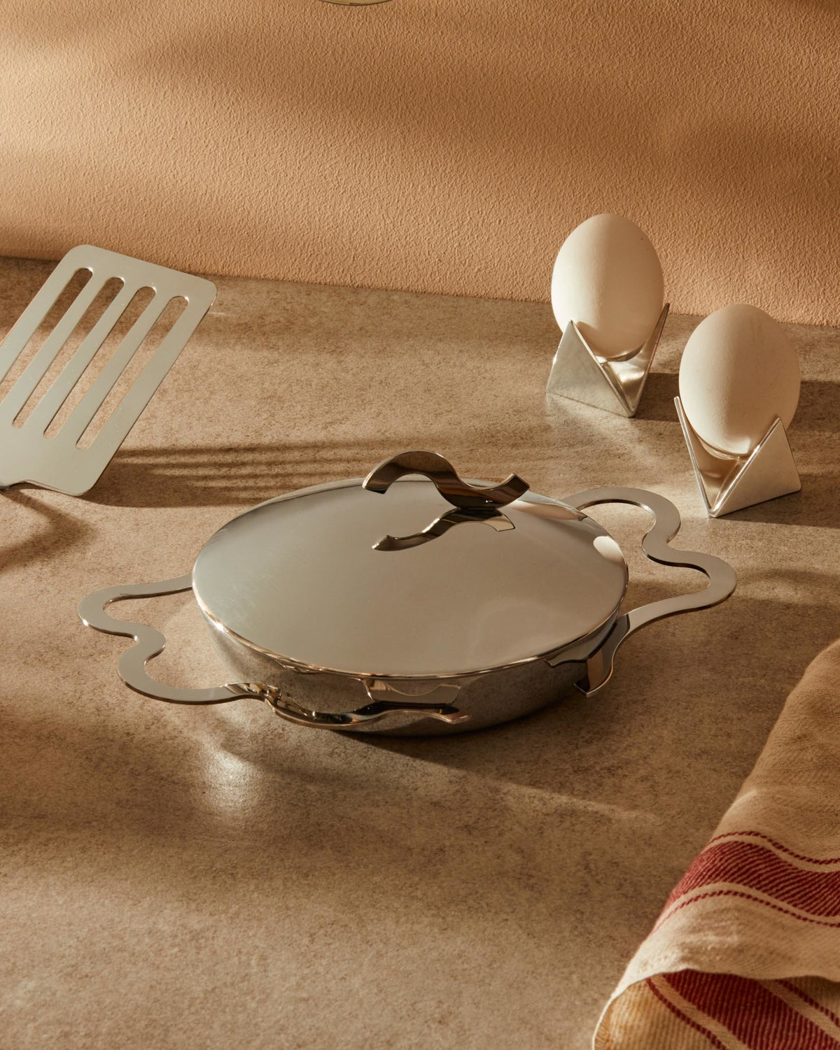 Silver metallic geometric egg cups by Alessi, holding white eggs on a speckled counter with a shiny pan, spatula, and striped towel.