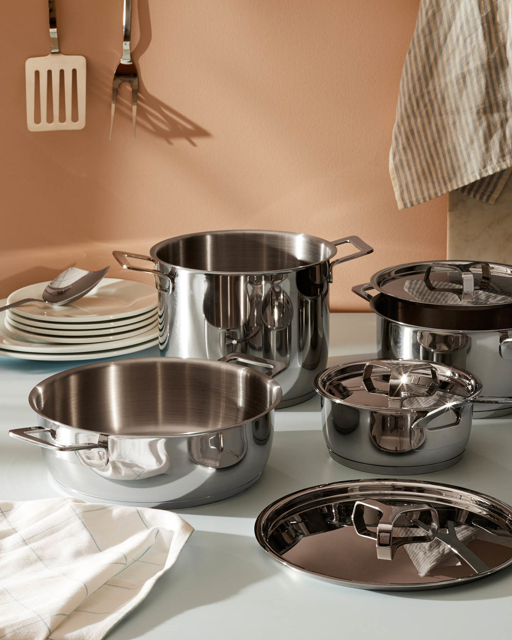 Shiny stainless steel pots and pans set by Alessi, including various sizes, displayed on a countertop with kitchen accessories.