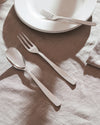 Shiny silver cutlery set by Alessi, with a spoon and two forks on a textured linen tablecloth, next to stacked white plates.