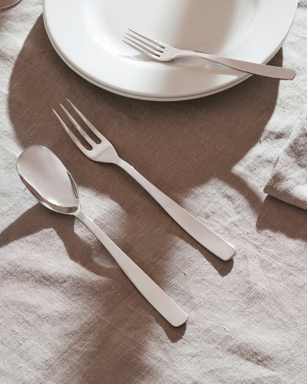 Shiny silver cutlery set by Alessi, with a spoon and two forks on a textured linen tablecloth, next to stacked white plates.