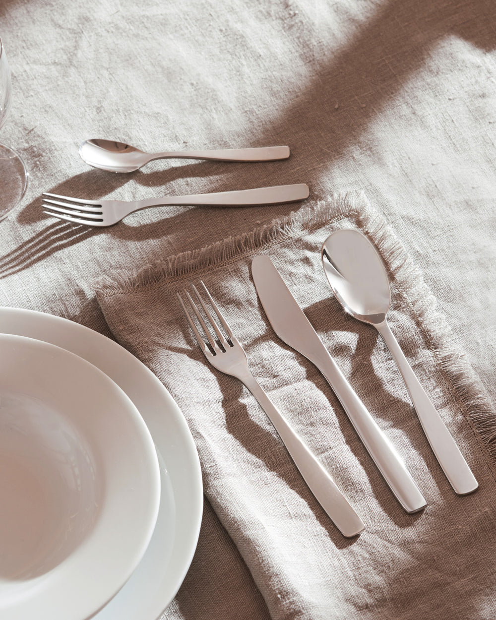 Sleek silver-toned knife, fork, and spoon set by Alessi, resting on a textured beige linen tablecloth.