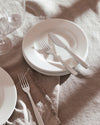 Silver stainless steel cutlery set by Alessi, showcasing a knife and two forks on stacked white plates with a frayed linen napkin.