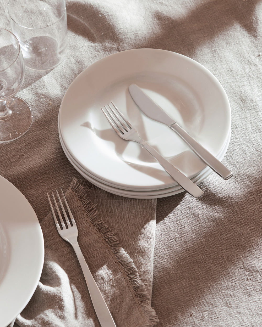Silver stainless steel cutlery set by Alessi, showcasing a knife and two forks on stacked white plates with a frayed linen napkin.