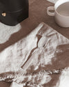 Silver spoon by Alessi on a light brown linen napkin with frayed edges, next to a black coffee maker and a white cup.