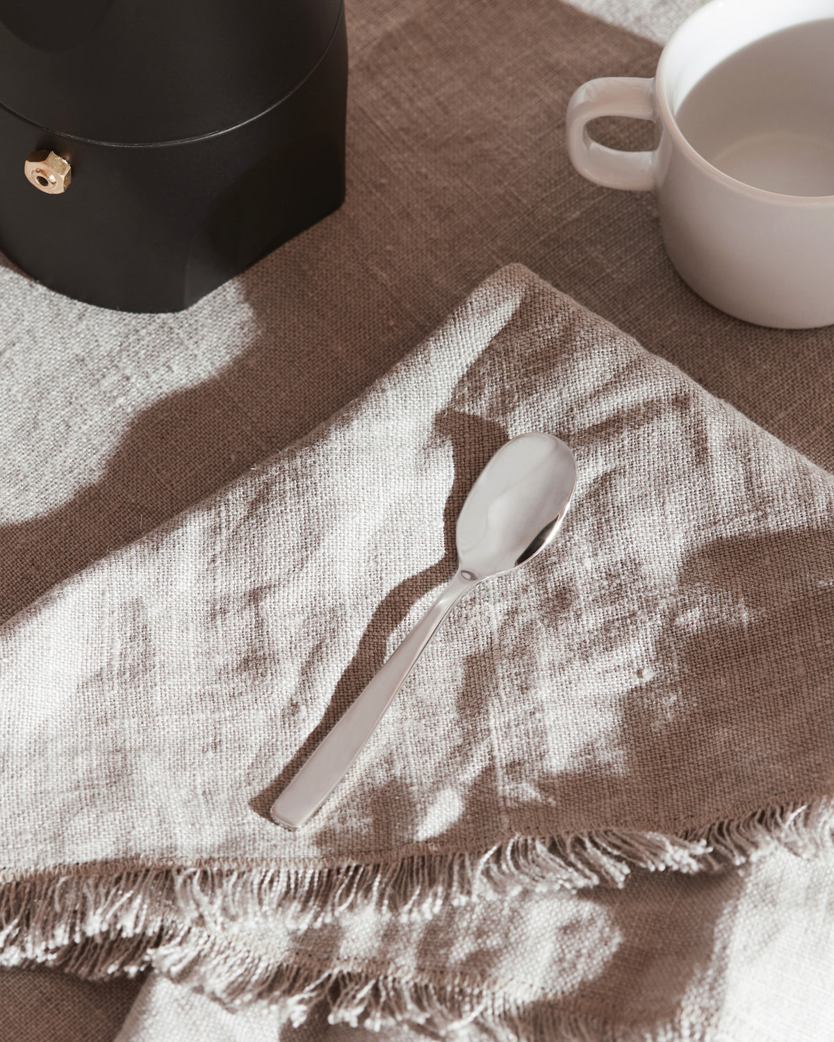 Silver spoon by Alessi on a light brown linen napkin with frayed edges, next to a black coffee maker and a white cup.