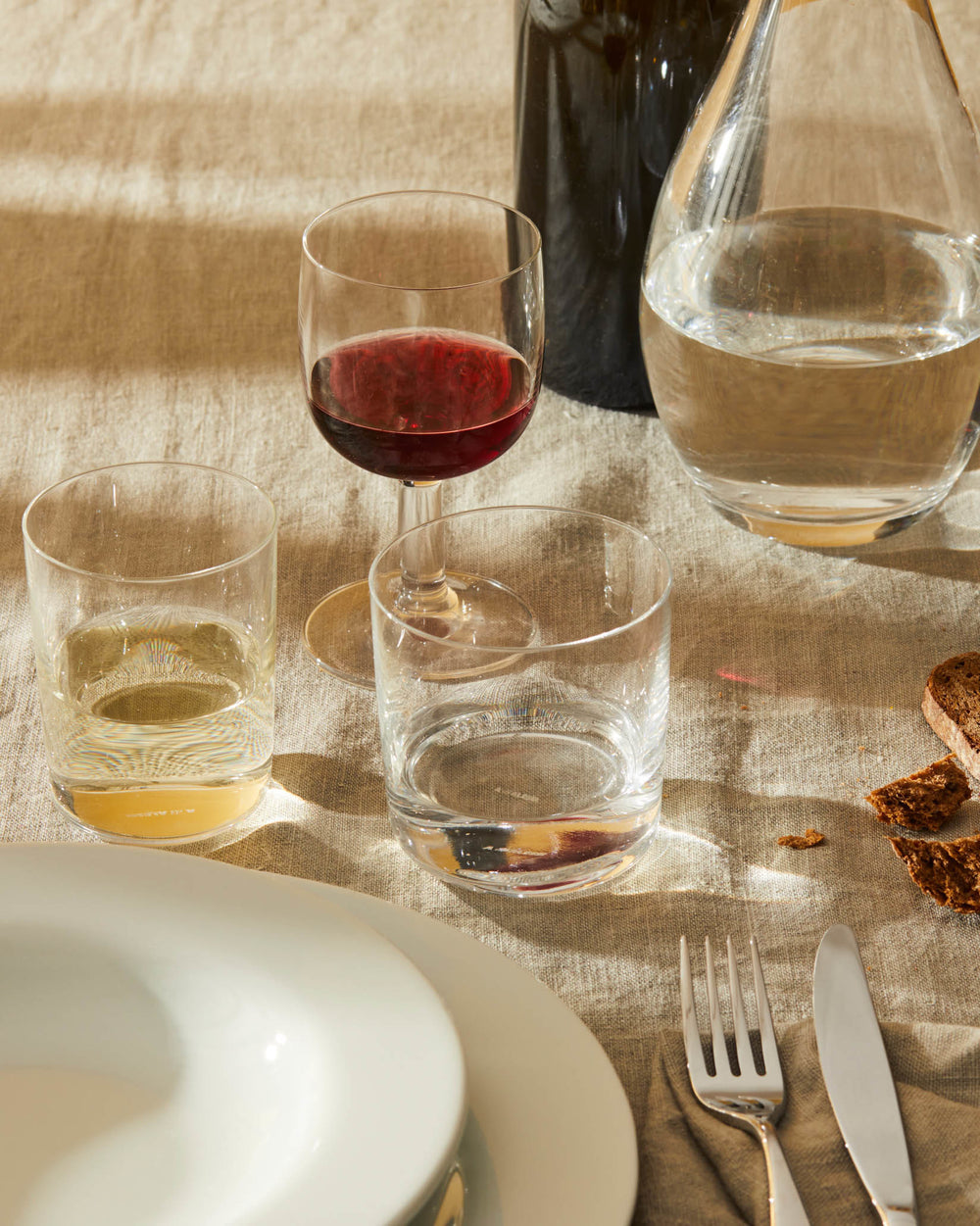 A collection of clear drinking glasses by Alessi, set on a rustic linen table with beverages and dinnerware.