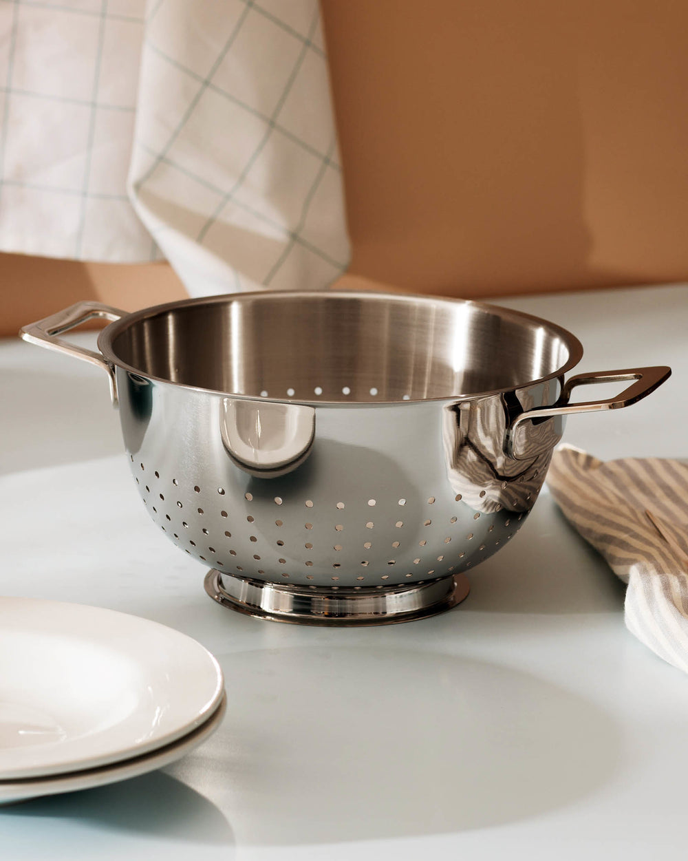 Polished stainless steel colander by Alessi with perforated bowl and two handles, on a light kitchen counter with white plates.