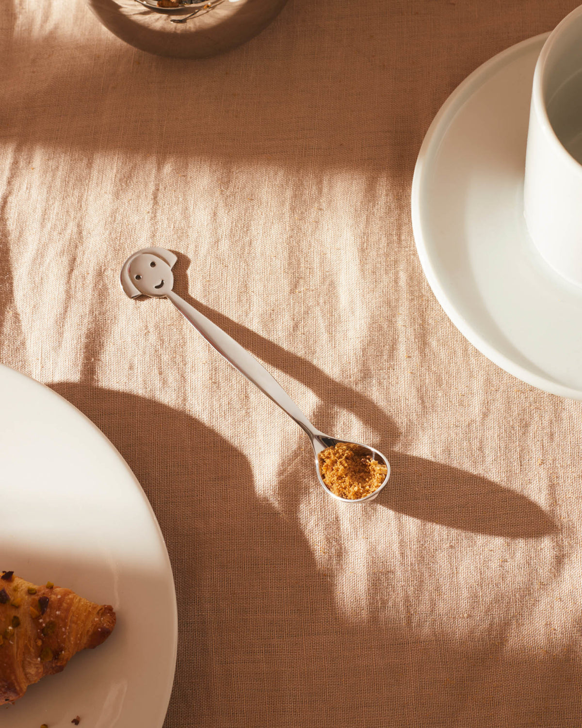 Silver-colored metal coffee spoon by Alessi with a smiling face handle, holding brown sugar on a beige linen tablecloth.