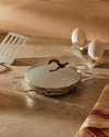 Polished stainless steel lidded egg pan by Alessi with a wavy base and decorative lid handle, next to eggs and a spatula.