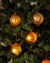 Golden Asinello Christmas ornaments by Alessi, with stylized girl, boy, cow, and pig faces, on a green tree.