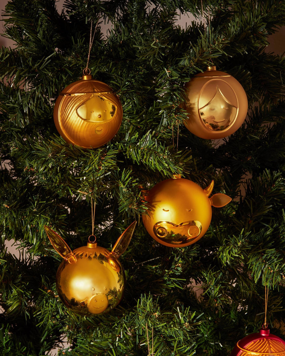 Golden Asinello Christmas ornaments by Alessi, with stylized girl, boy, cow, and pig faces, on a green tree.