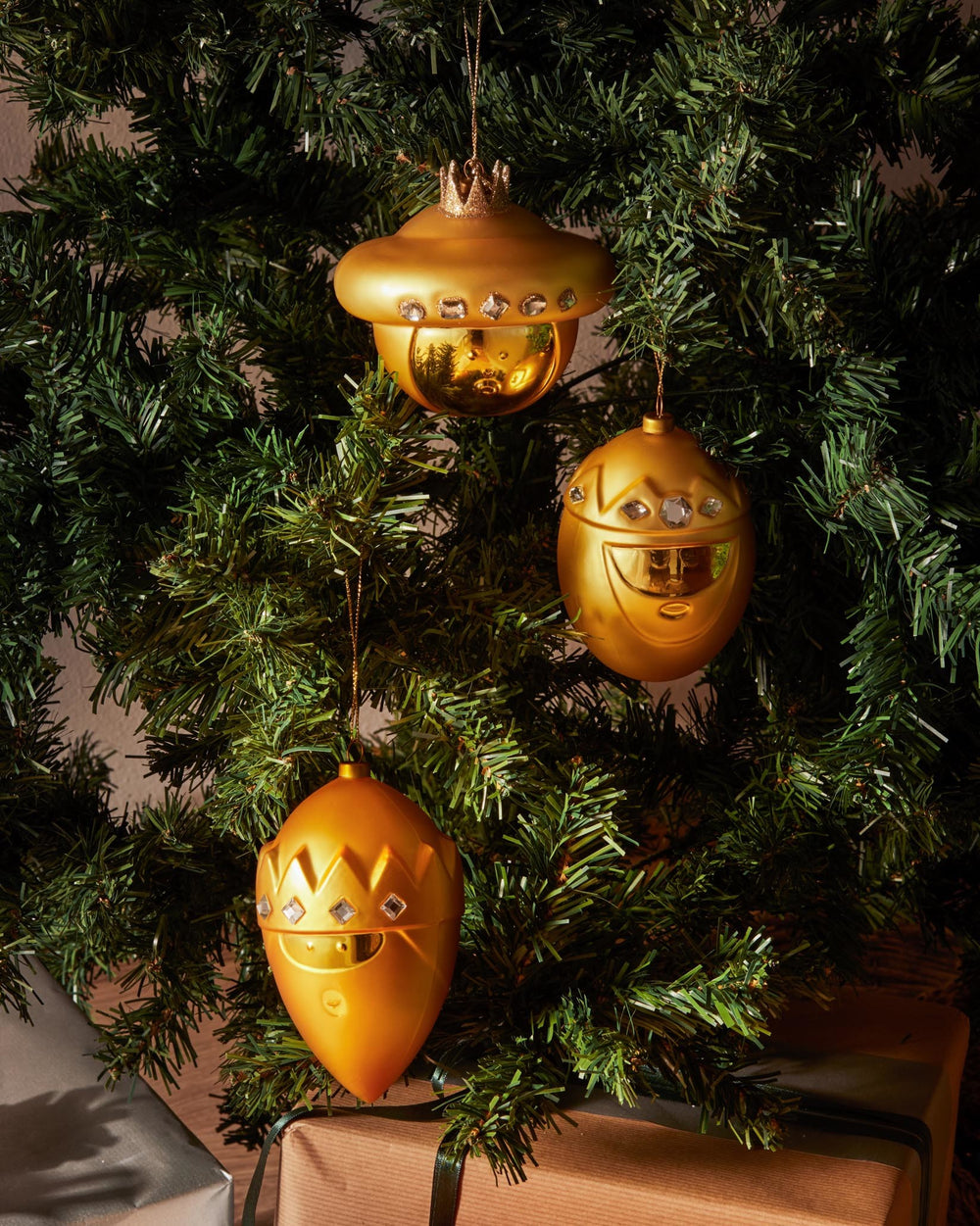 Golden crown-shaped Christmas ornament by Alessi with a glitter crown, clear faceted gems, and reflective bottom, hanging on a dark green Christmas tree.