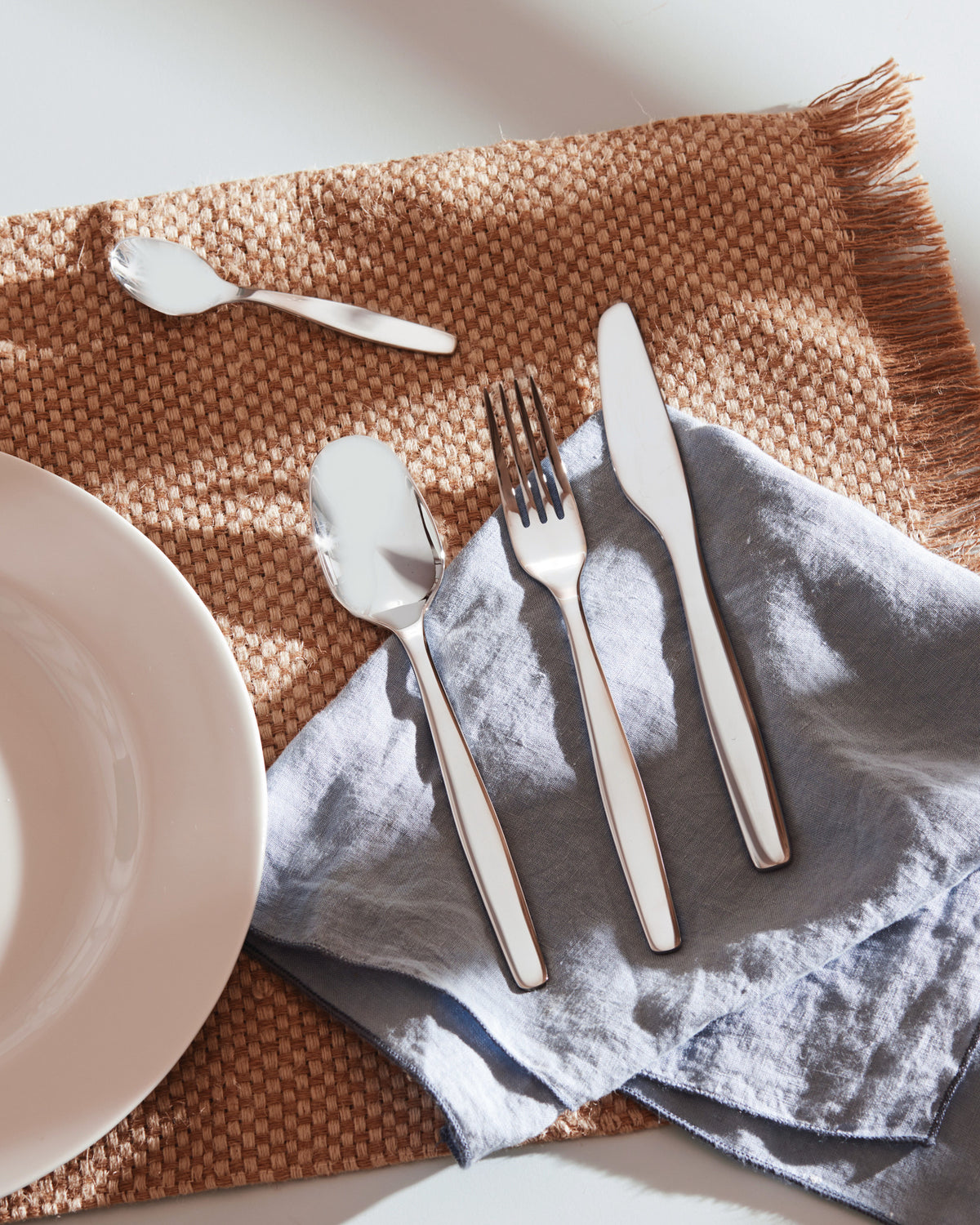 Itsumo silver cutlery set by Alessi on a brown woven placemat with a blue linen napkin and white plate.