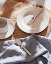 Itsumo silver-tone cutlery by Alessi, with a knife on a white plate and a spoon on a blue linen napkin, cast in sunlight.