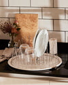 Silver wire dish rack by Alessi, round with a cream tray, holding clean dishes on a black countertop.