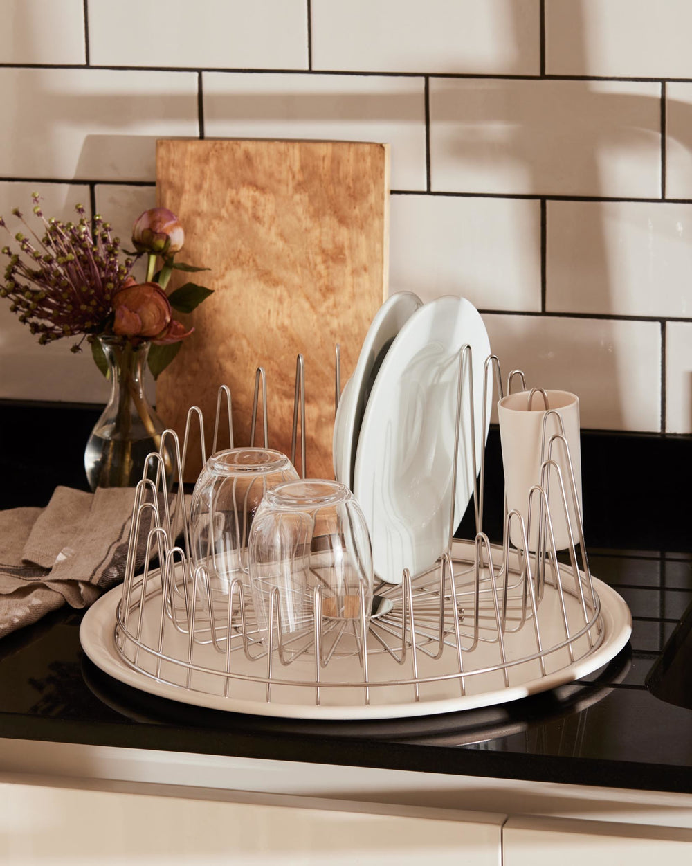 Silver wire dish rack by Alessi, round with a cream tray, holding clean dishes on a black countertop.