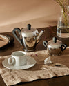 Polished chrome tea set with black handles by Alessi, featuring a teapot, sugar bowl, and white teacup.