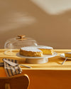 Clear glass cake dome with a natural wood knob and white ceramic stand by Alessi, displaying a sliced cake on a yellow tablecloth.
