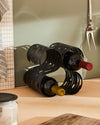 Black metal wavy S-shaped wine rack by Alessi holding two bottles on a wooden surface.
