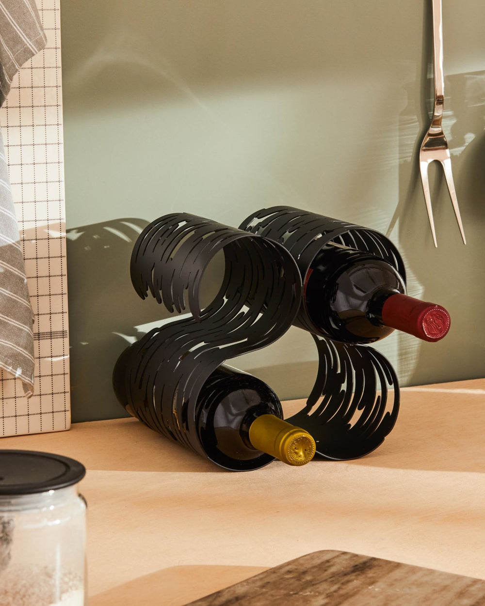 Black metal wavy S-shaped wine rack by Alessi holding two bottles on a wooden surface.