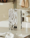 Silver metallic paper towel holder by Alessi featuring an organic cut-out design, holding a white roll in a kitchen.