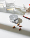 Silver and clear round pill organizer by Alessi, on a white table with water glasses and scattered pills.