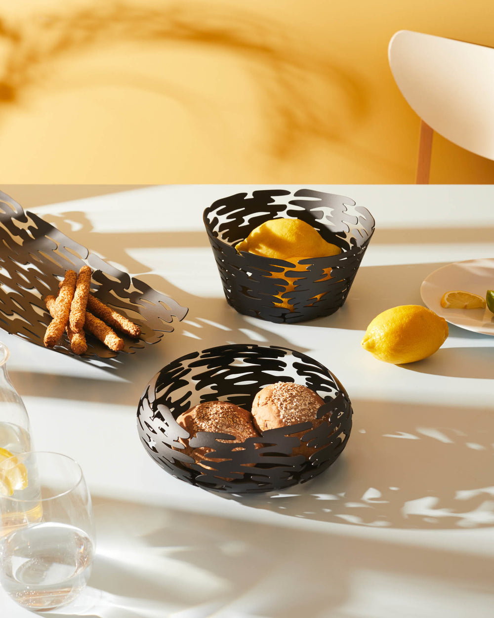 Black metal basket by Alessi with an organic bark-like cut-out pattern, displaying lemons and bread.