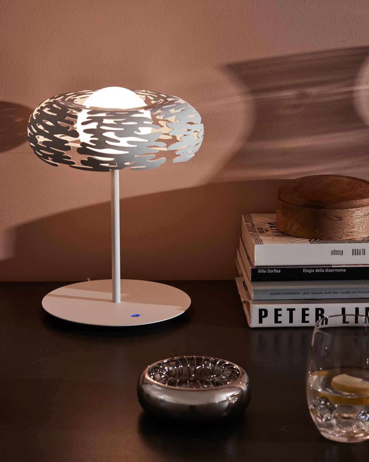 White table lamp by Alessi with an intricate, organic-patterned shade, casting shadows on a wall, styled on a dark surface.