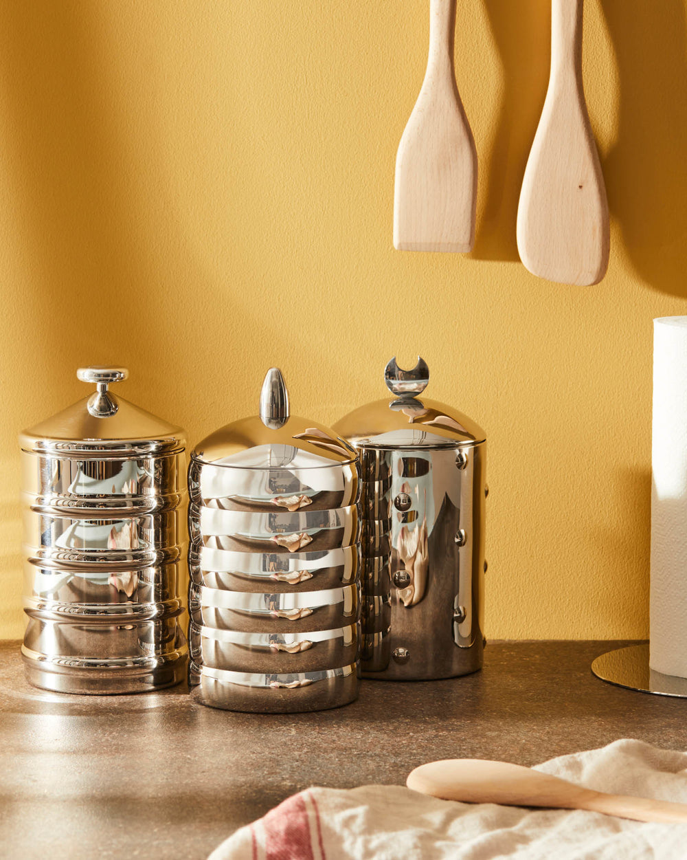 Three polished metal jars by Alessi, each with a distinctive finial and ribbed details, on a countertop.